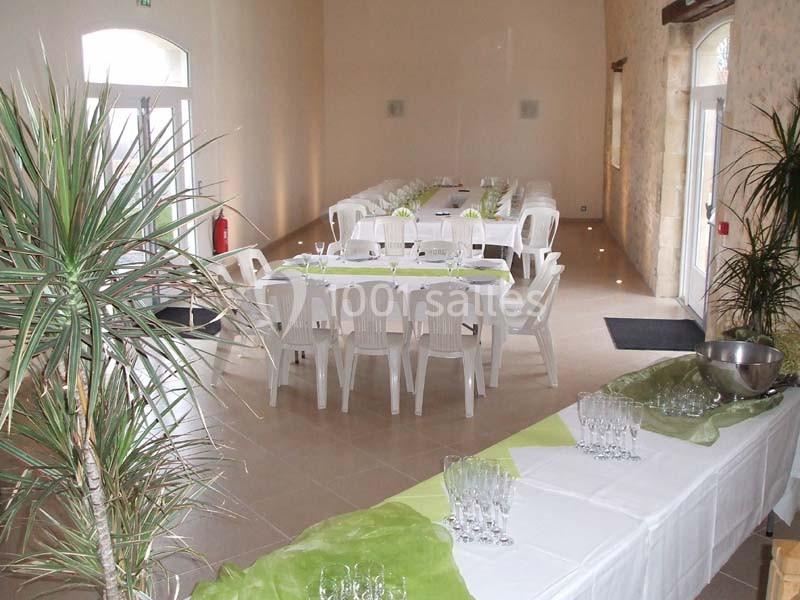 Salle lumineuse aménagée pour un événement, avec tables décorées de nappes blanches et vertes, et plantes en décoration.
