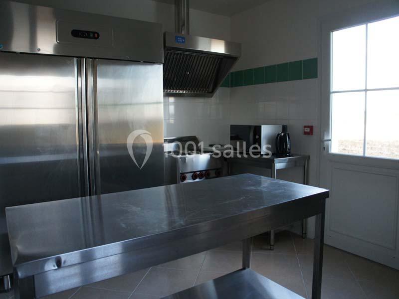 Cuisine professionnelle avec réfrigérateur en inox, hotte, plan de travail métallique, micro-ondes et fenêtre lumineuse.