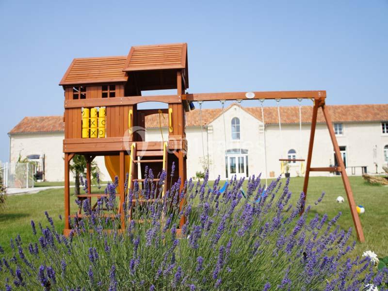 Structure en bois avec toboggan et balançoire dans un jardin, entourée de lavandes et d'un bâtiment en arrière-plan.
