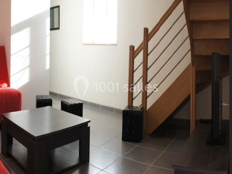 Salon lumineux avec escalier en bois, table basse sombre, fauteuil rouge et sol en carrelage gris.