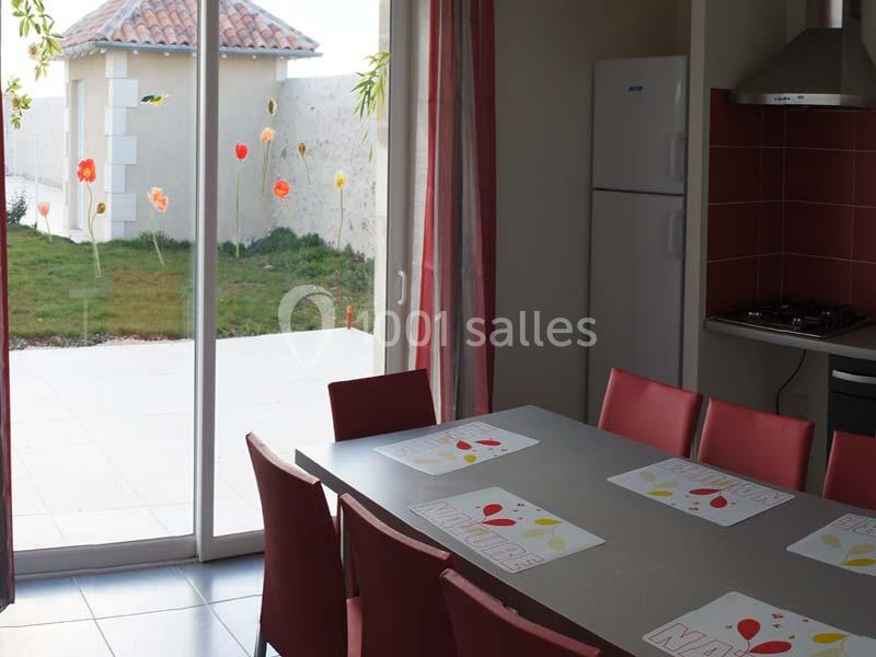 Salle à manger lumineuse avec table, chaises rouges, cuisine équipée et vue sur un jardin par une baie vitrée.
