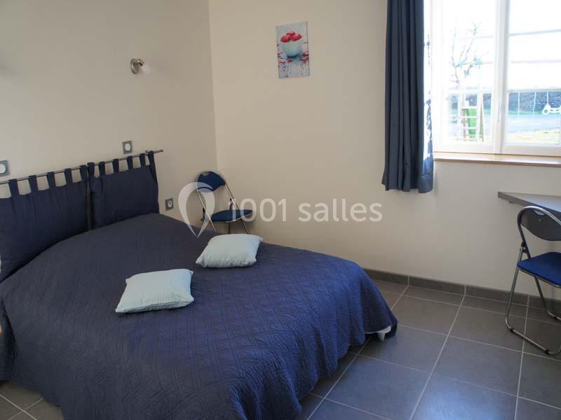 Chambre lumineuse avec un lit double, couvre-lit bleu, deux coussins, une fenêtre et un bureau avec chaise.