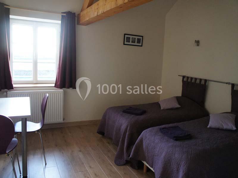 Chambre avec deux lits simples, couvre-lits violets, table, chaises et fenêtre laissant entrer la lumière naturelle.