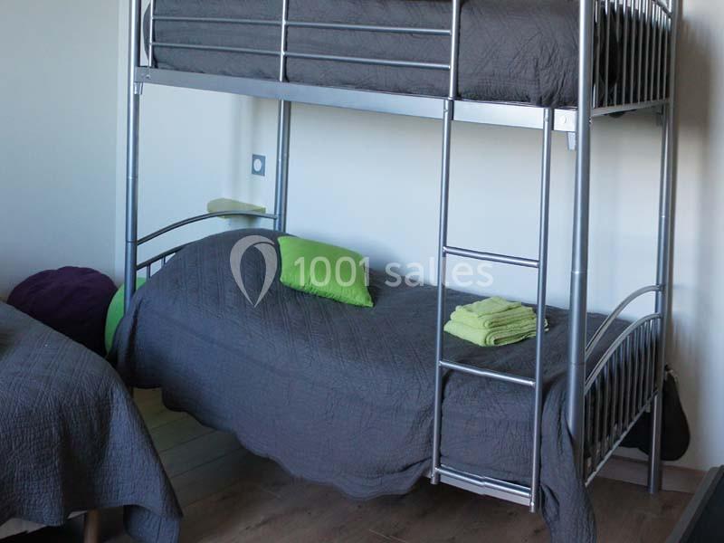 Lit superposé en métal avec draps gris, coussins verts et une pile de serviettes sur le lit du bas.