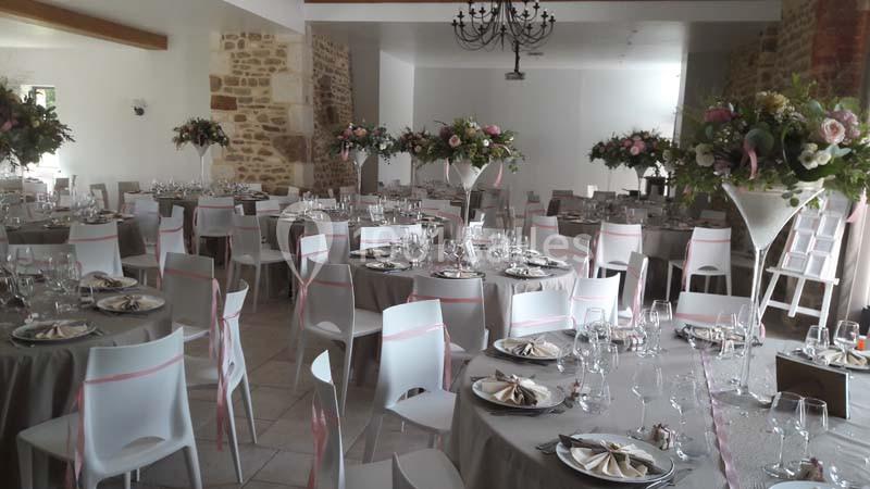 Salle de réception décorée avec des tables rondes, nappes grises, chaises blanches et arrangements floraux élégants.
