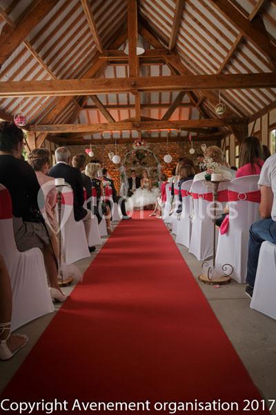 Cérémonie de mariage dans une grange décorée, avec invités assis de part et d'autre d'une allée rouge.