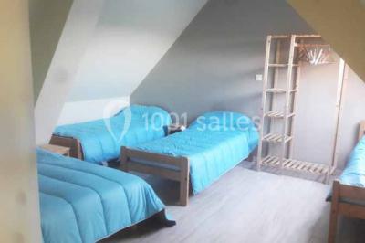 Chambre mansardée avec quatre lits simples, couvertures bleues et étagère en bois clair contre un mur gris.