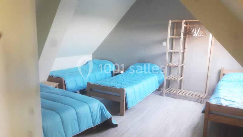 Chambre mansardée avec quatre lits simples, couvertures bleues et étagère en bois clair contre un mur gris.