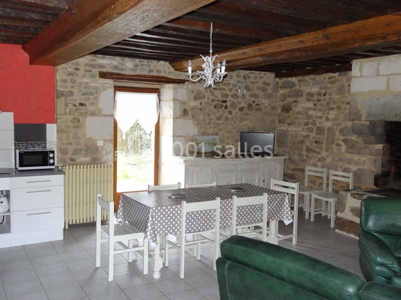 Salle à manger rustique avec murs en pierre, table rectangulaire, chaises blanches, cuisine équipée et cheminée en pierre.