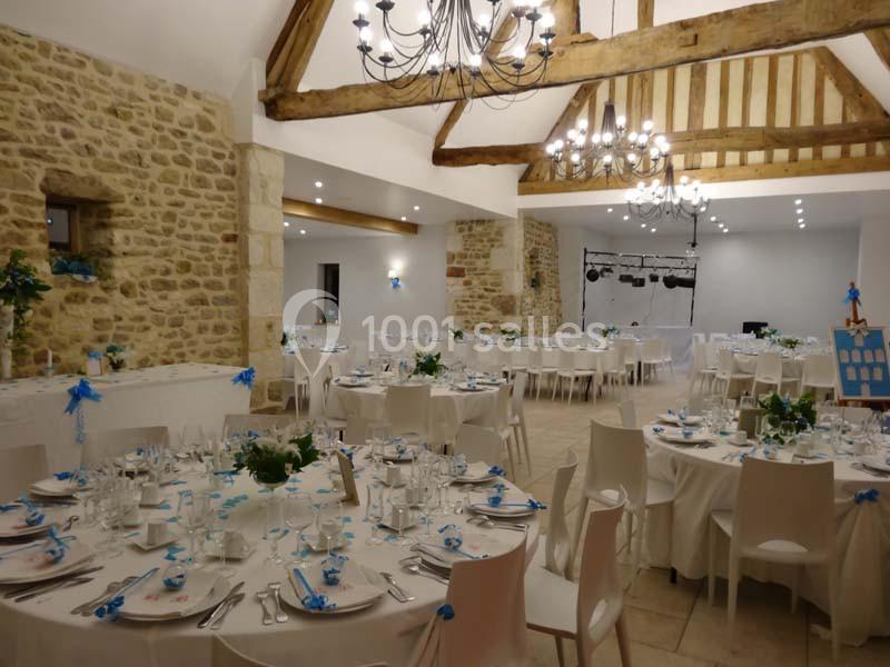 Salle de réception décorée avec des tables rondes dressées, des chaises blanches et des poutres apparentes au plafond.