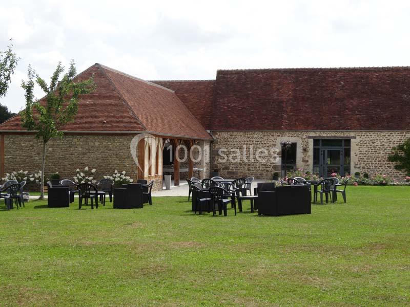 Bâtiment en pierre avec toit en tuiles, terrasse aménagée de chaises et tables noires sur une pelouse.