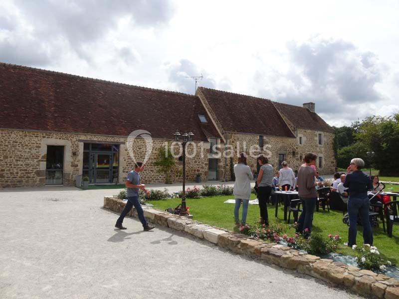 Des personnes discutent et se détendent autour de tables dans une cour devant des bâtiments en pierre sous un ciel nuageux.