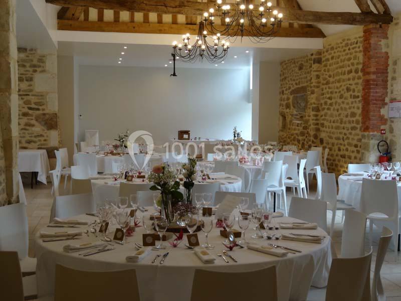 Salle de réception lumineuse avec tables rondes dressées, décorée de fleurs et éclairée par un lustre en fer forgé.