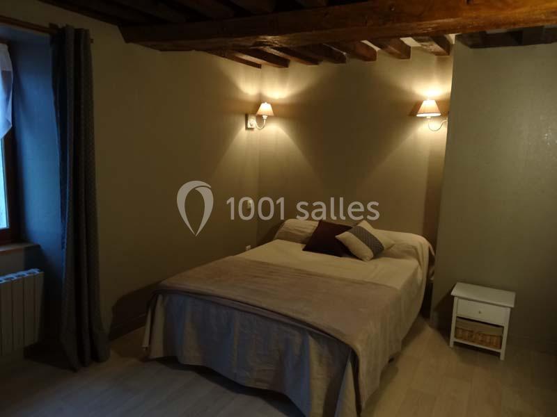 Chambre avec un lit double, deux appliques murales allumées, un plafond avec poutres apparentes et une fenêtre avec rideaux.