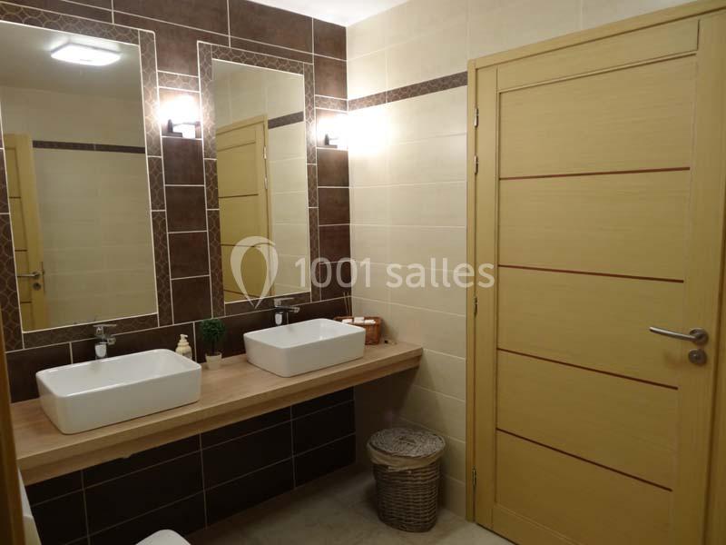 Salle de bain moderne avec deux lavabos, miroirs éclairés, murs carrelés marron et beige, et une porte en bois clair.