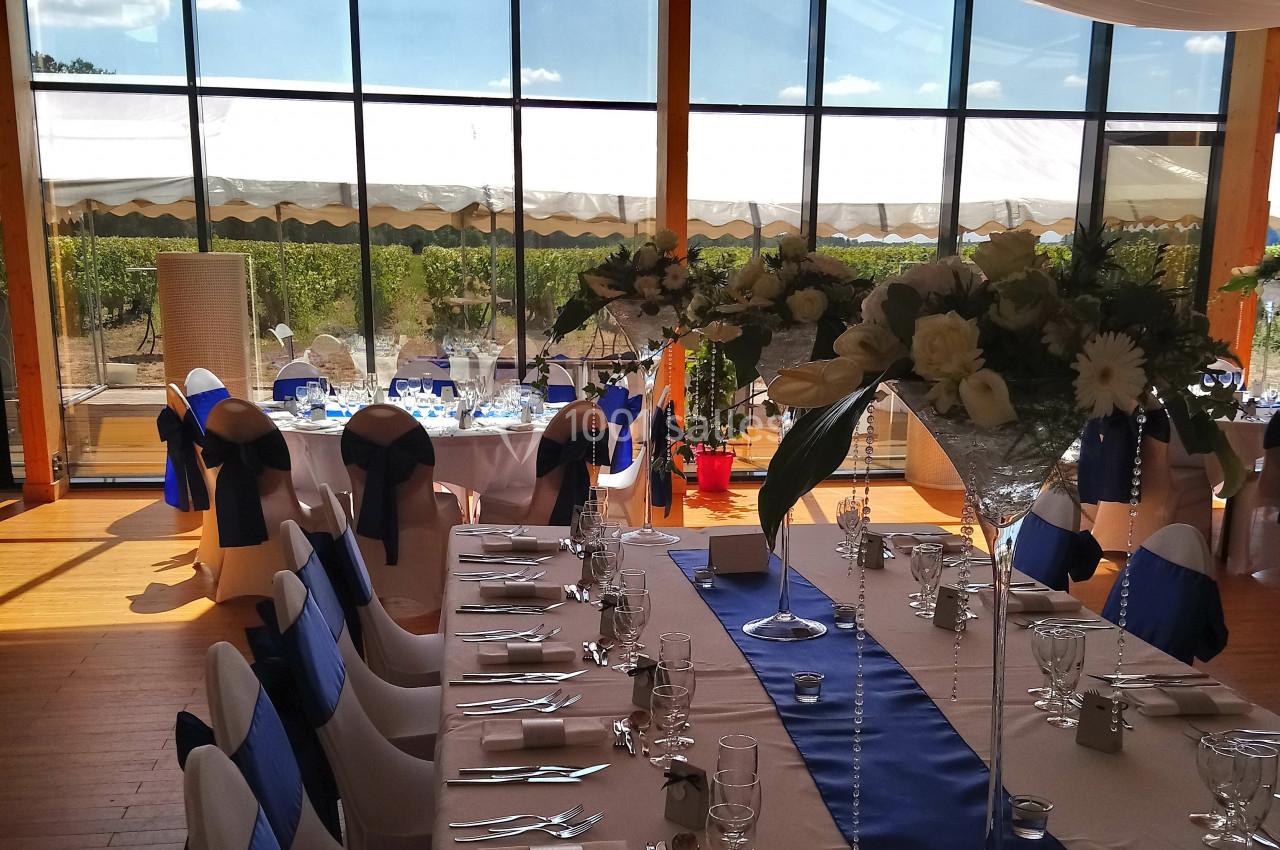 Salle de réception lumineuse avec tables décorées de nappes blanches et bleues, et vue sur un paysage extérieur ensoleillé.
