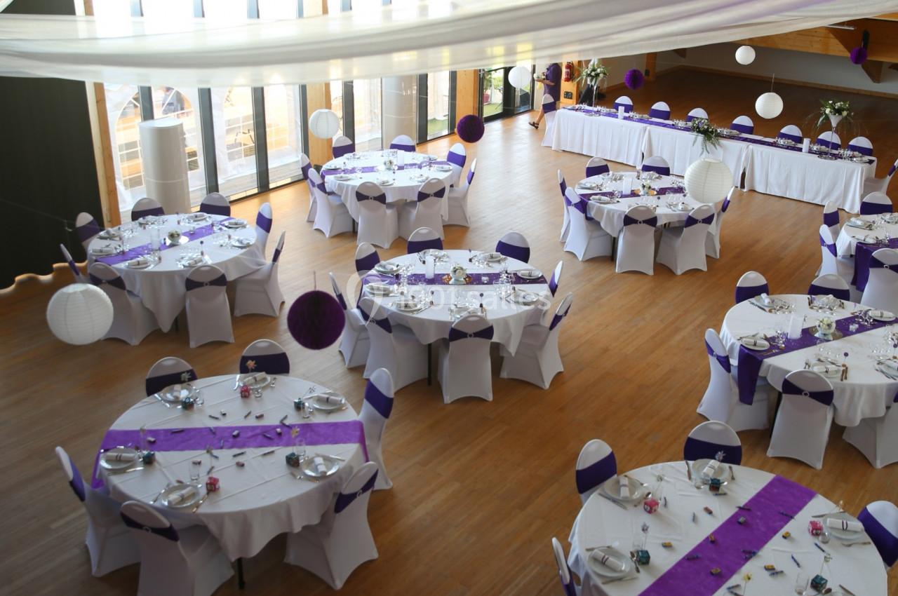 Salle de réception décorée avec des tables rondes dressées, nappes blanches et chemins de table violets.