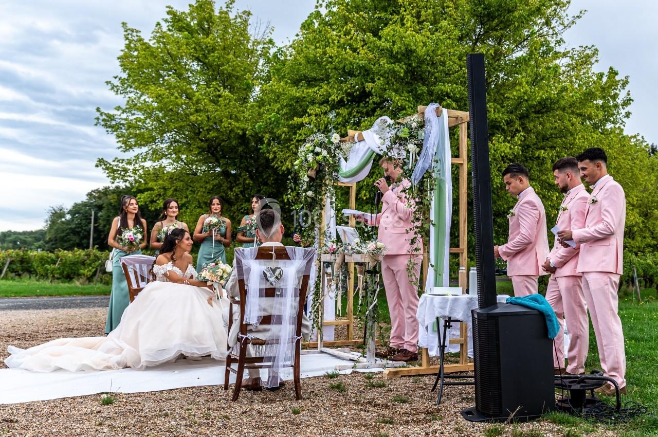 Cérémonie de mariage en plein air avec des mariés assis, un officiant et des invités en tenues élégantes.