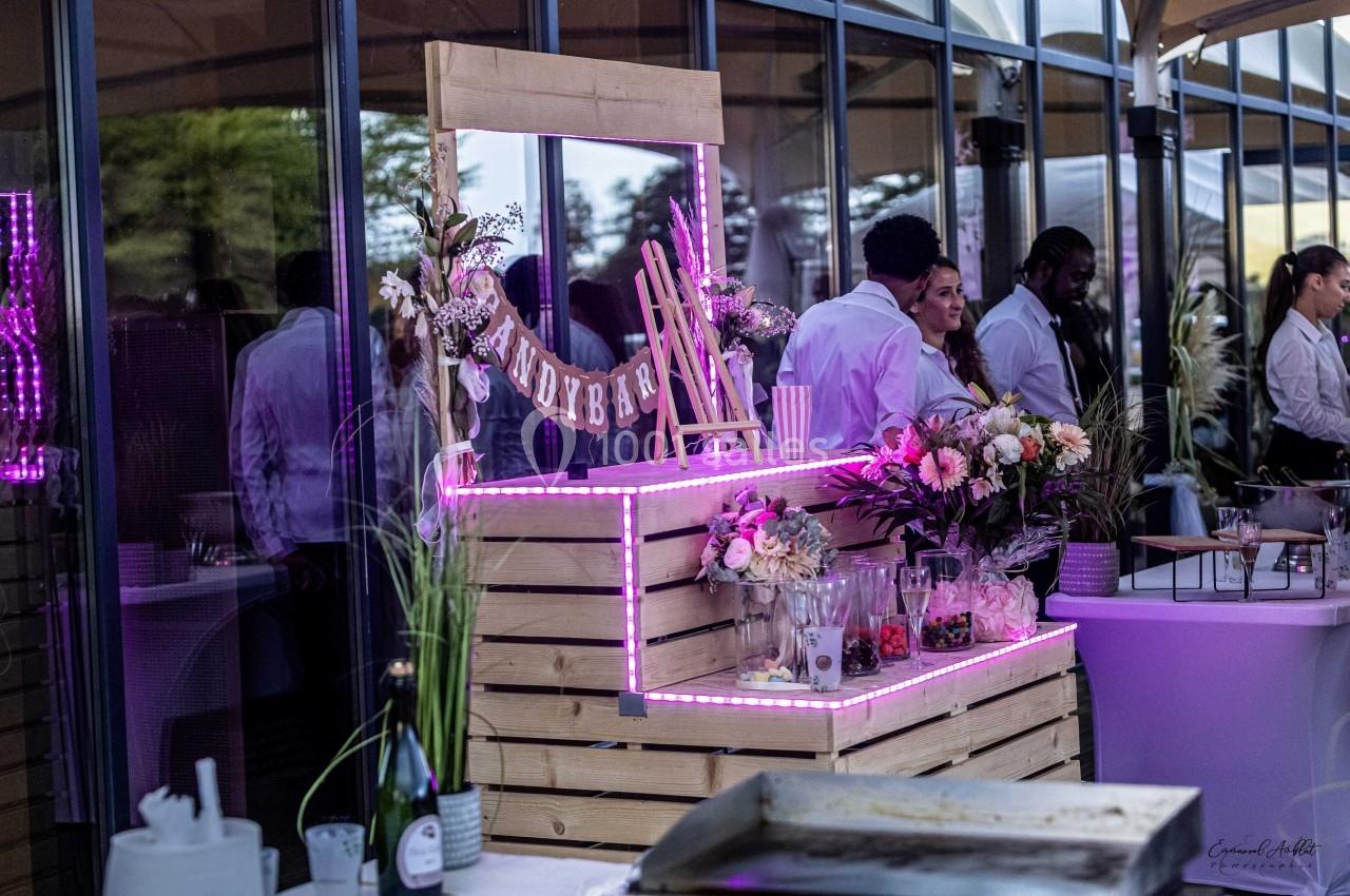 Stand de bar lumineux en bois décoré de fleurs, avec des verres et des ingrédients, lors d'un événement en extérieur.