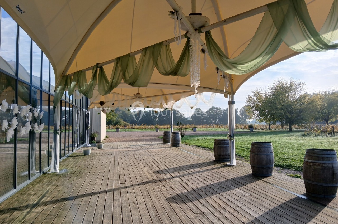 Terrasse en bois couverte par une toile beige, décorée de voilages verts et de barriques, avec vue sur un paysage champêtre.