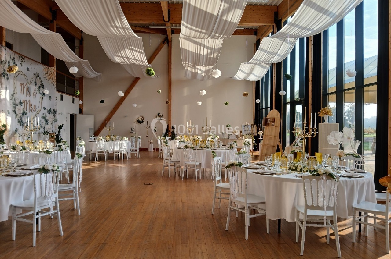 Salle de réception décorée pour un mariage, avec tables dressées, nappes blanches et voilages suspendus au plafond.