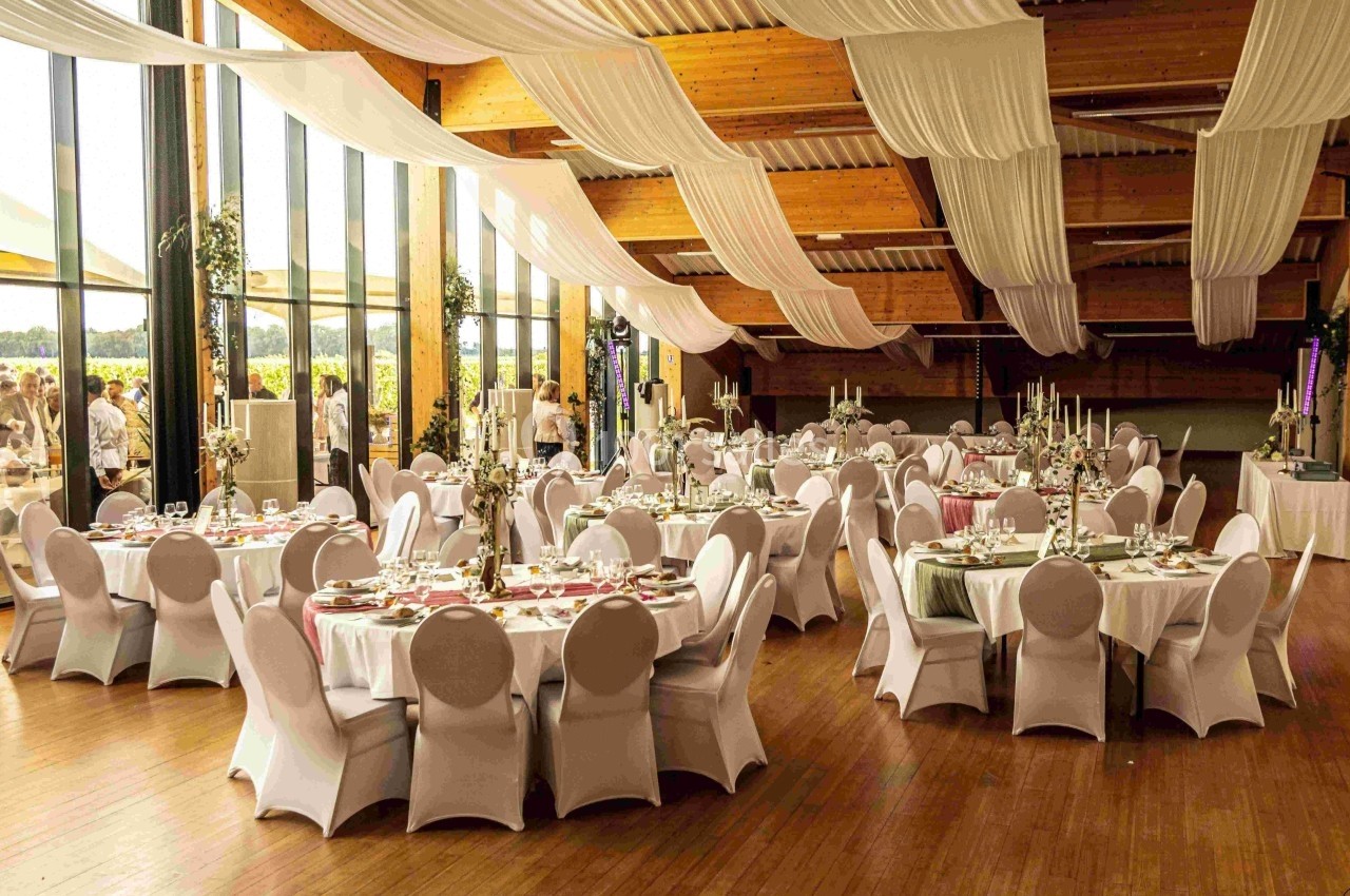 Salle de réception décorée avec des tables rondes dressées, nappes blanches et plafond orné de drapés.