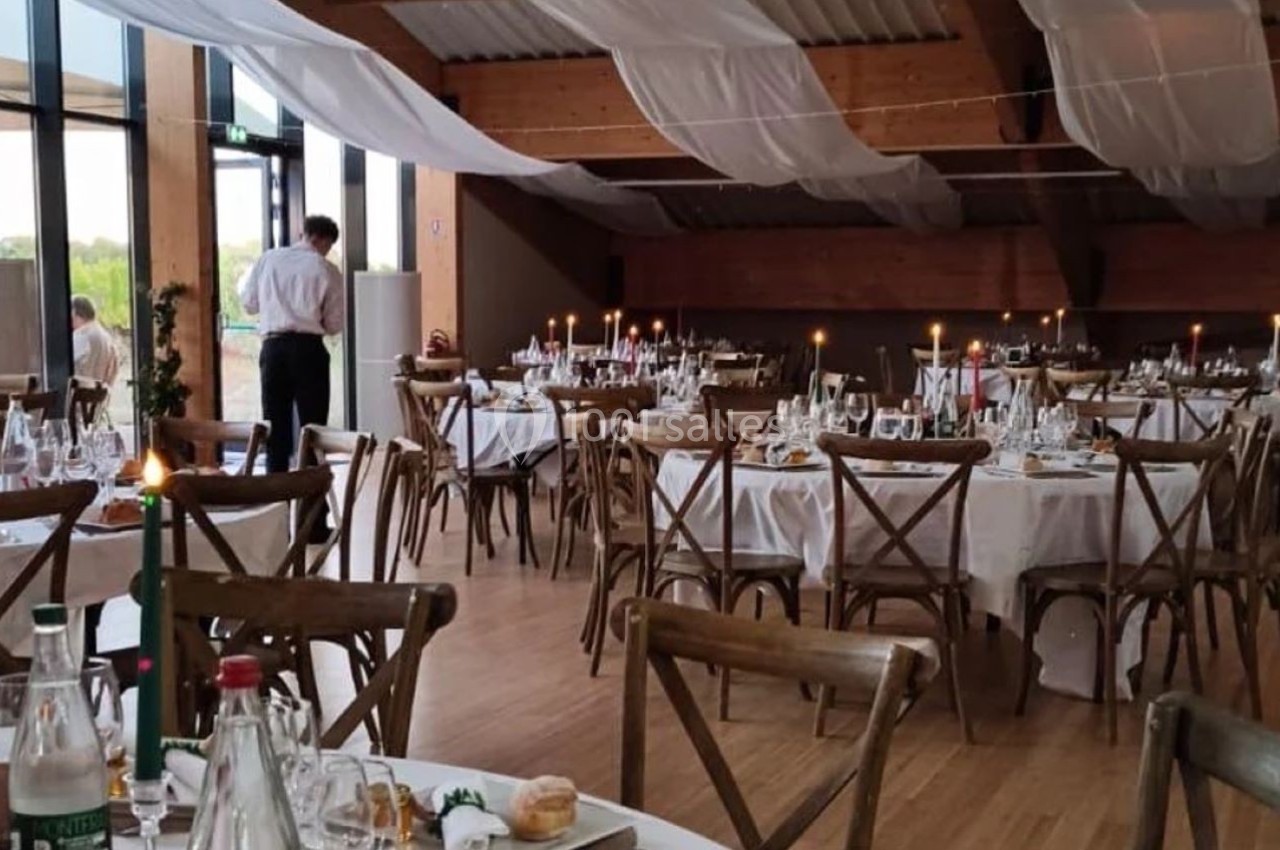 Salle de réception décorée avec des tables rondes dressées, bougies allumées et nappes blanches, sous un plafond en bois.