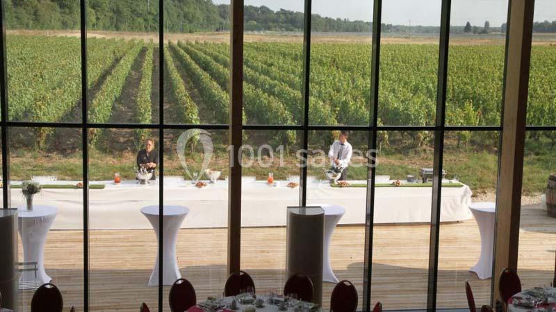 Vue sur un vignoble à travers de grandes baies vitrées, avec une table dressée en extérieur pour un événement.