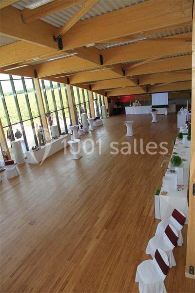 Grande salle lumineuse avec parquet en bois, tables hautes et chaises blanches, baies vitrées donnant sur un extérieur…
