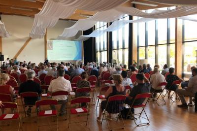 Salle lumineuse accueillant un salon avec des stands, des visiteurs et des tables exposant divers produits et services.