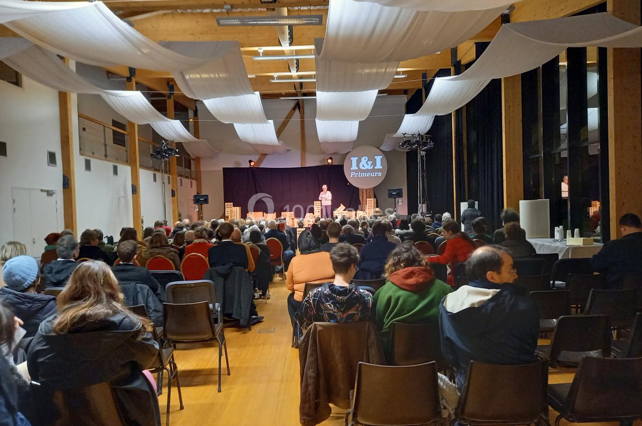 Salle remplie de spectateurs assis, écoutant un intervenant sur une scène éclairée dans un cadre avec poutres en bois.