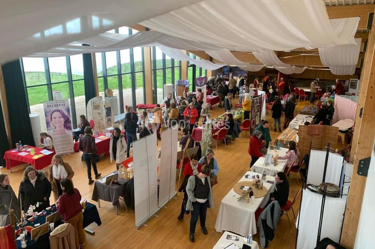 Salle lumineuse accueillant un salon avec des stands, des visiteurs et des tables exposant divers produits et services.