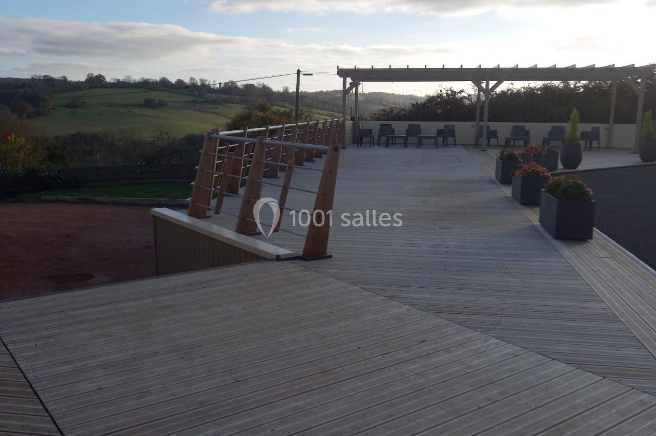 Terrasse en bois avec rampe d'accès, pergola et vue dégagée sur un paysage rural sous un ciel partiellement nuageux.