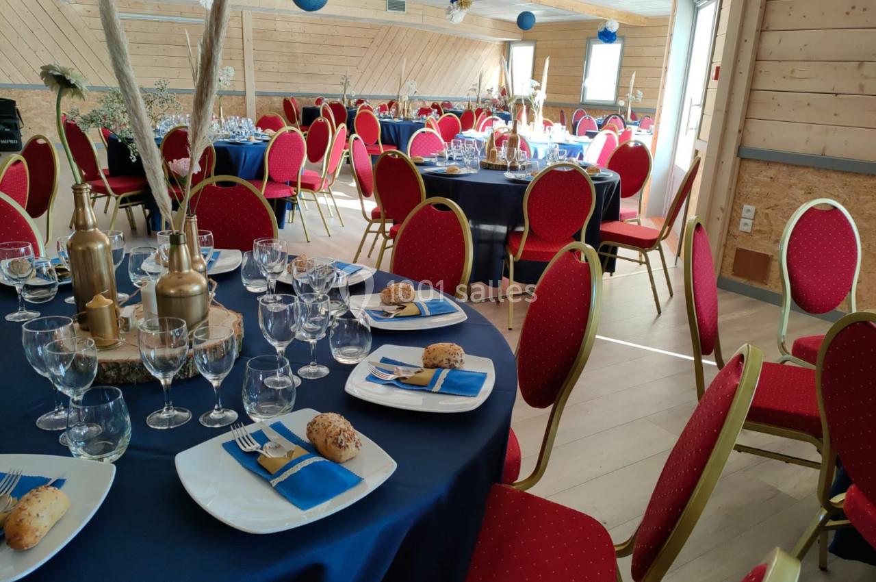 Salle de réception décorée avec des tables rondes dressées, nappes bleues, chaises rouges et éléments décoratifs dorés.