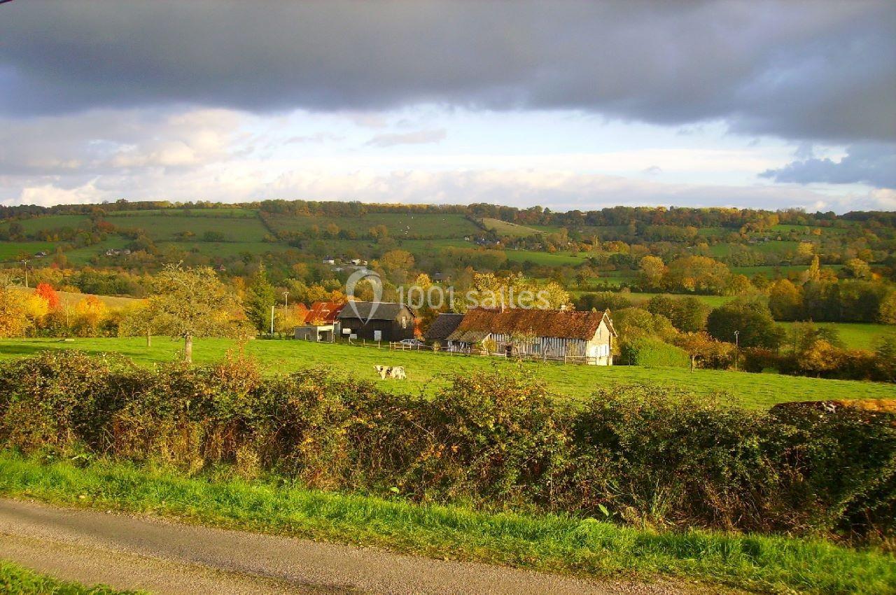Paysage rural avec une ferme traditionnelle entourée de prairies verdoyantes et de collines sous un ciel nuageux.