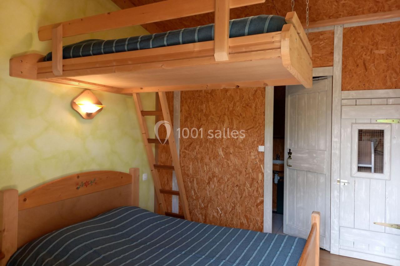 Chambre en bois avec lit double, mezzanine avec matelas, échelle en bois et porte blanche sur un mur texturé.