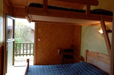 Chambre simple avec un lit double, une table en bois, une chaise et une lumière murale, donnant sur une porte vitrée.