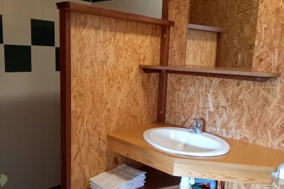 Chambre simple avec un lit double, une table en bois, une chaise et une lumière murale, donnant sur une porte vitrée.