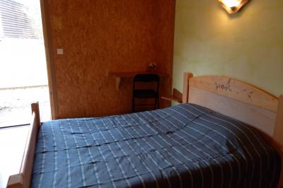 Chambre simple avec un lit double, une table en bois, une chaise et une lumière murale, donnant sur une porte vitrée.