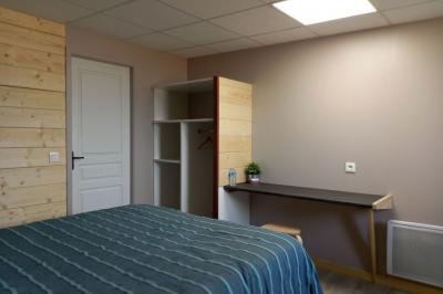 Chambre simple avec un lit double, une table en bois, une chaise et une lumière murale, donnant sur une porte vitrée.