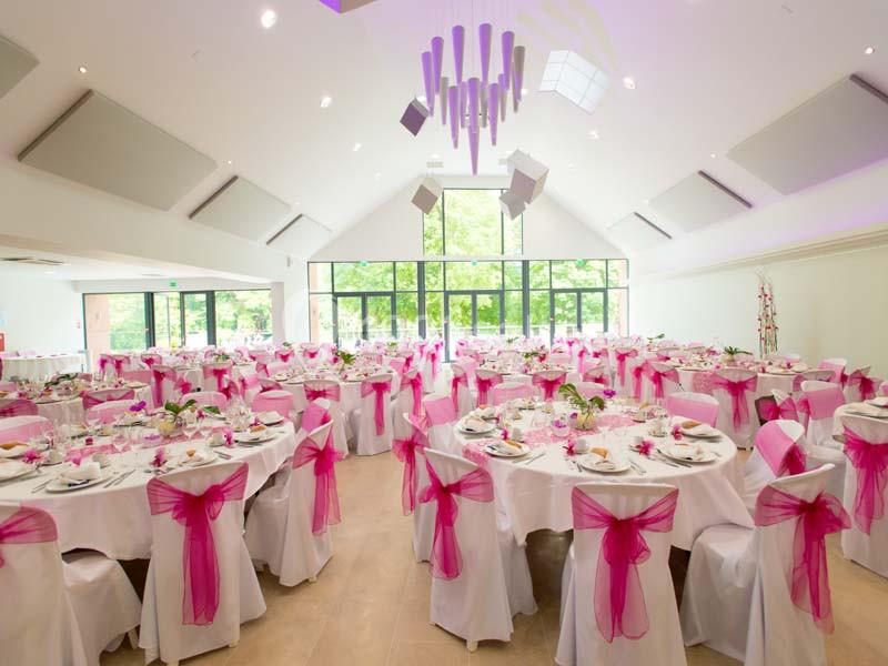 Salle de réception lumineuse décorée de nappes blanches et de nœuds roses sur les chaises, tables dressées pour un événement.