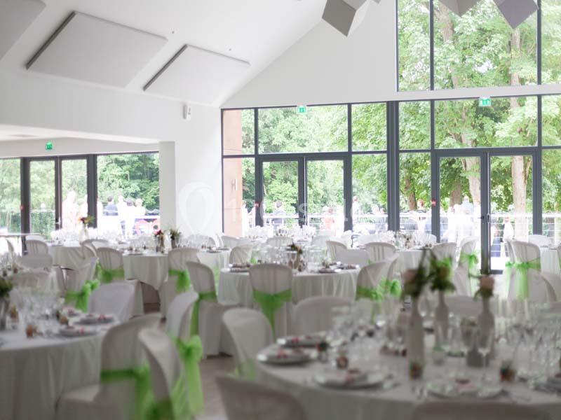 Salle de réception lumineuse avec tables décorées de nappes blanches et rubans verts, donnant sur un jardin verdoyant.