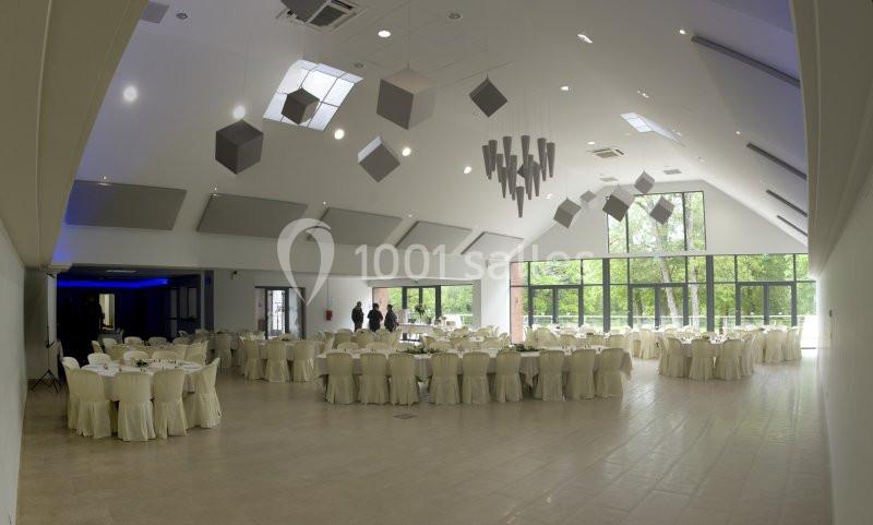 Salle de réception lumineuse avec tables rondes dressées, chaises couvertes et grandes baies vitrées donnant sur un espace…