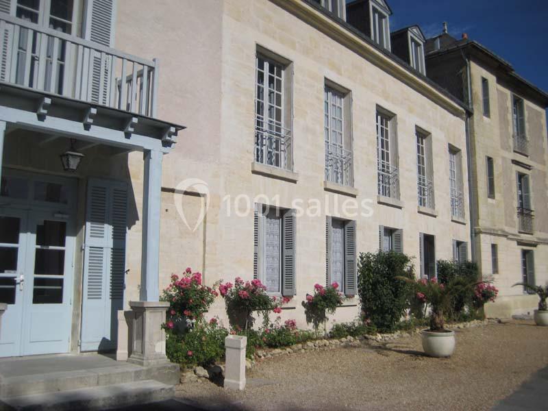 Façade d'un bâtiment en pierre claire avec volets bleus, jardinières fleuries et allée en gravier.