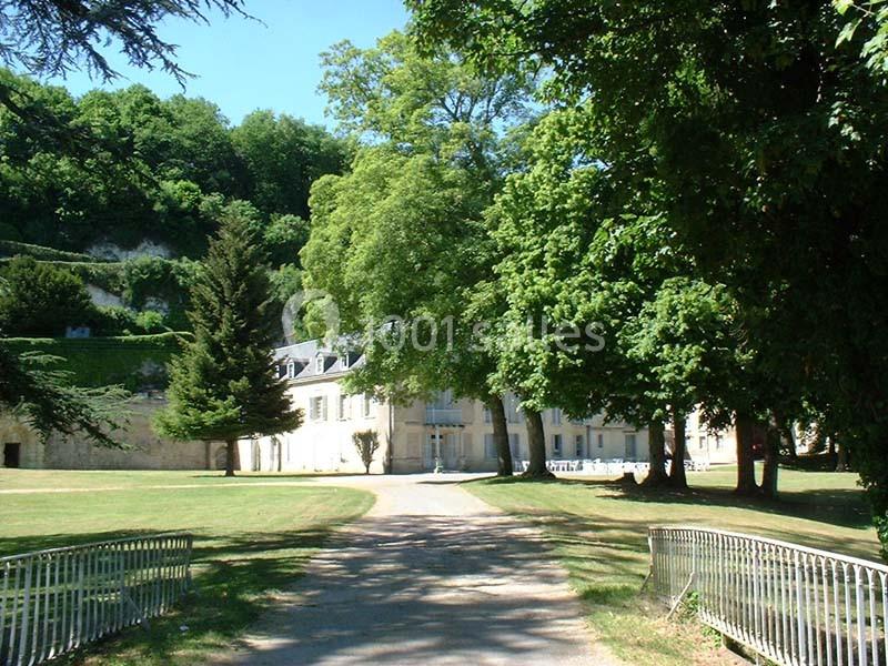 Allée bordée d'arbres menant à un bâtiment ancien entouré de verdure, par une journée ensoleillée.