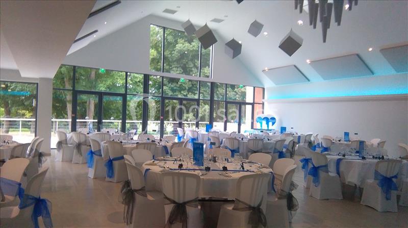 Salle de réception lumineuse avec des tables rondes dressées, décorées de nappes blanches et de nœuds bleus.