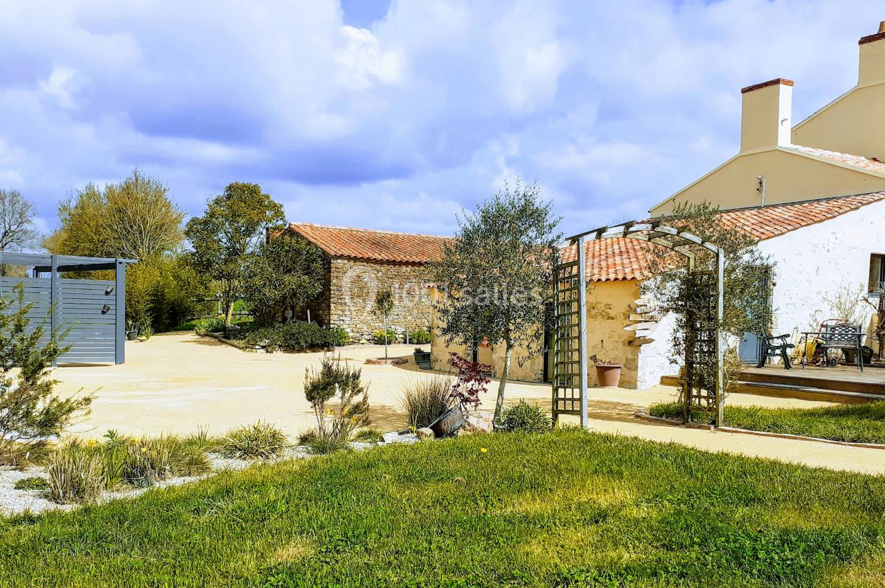 Cour d'une maison en pierre avec jardin, terrasse, pergola et ciel partiellement nuageux.