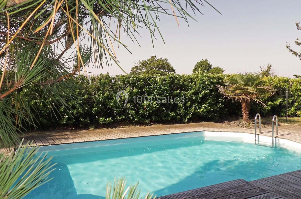 Piscine extérieure entourée de végétation, avec une terrasse en bois et un ciel dégagé en arrière-plan.