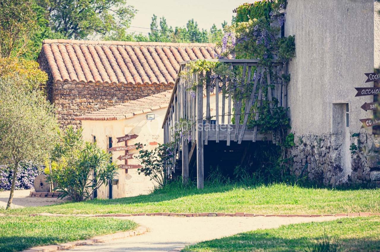 Chemin bordé de verdure menant à des bâtiments en pierre avec une terrasse en bois et des panneaux directionnels.