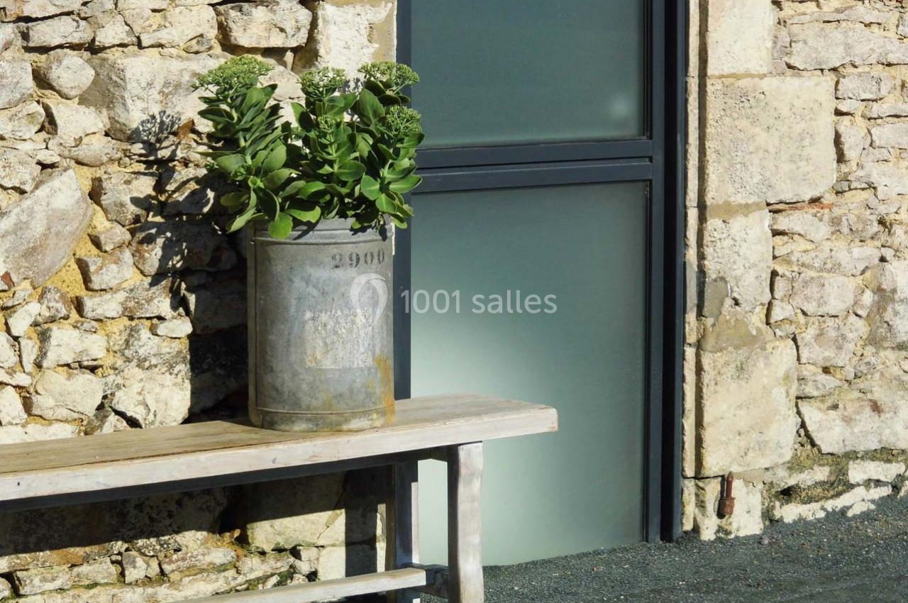 Un pot en métal contenant des plantes vertes posé sur un banc en bois devant un mur en pierre et une porte vitrée.